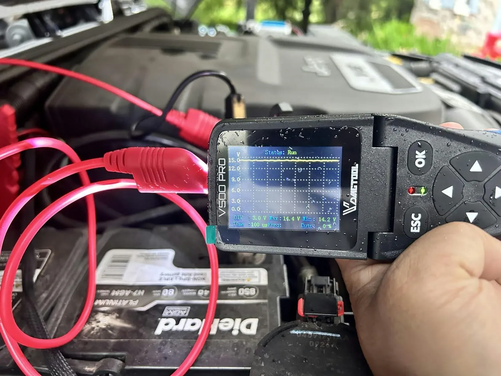 Handheld automotive circuit diagnostic tester connected to a car battery, showing a live voltage graph on its display with red test leads in an engine bay.